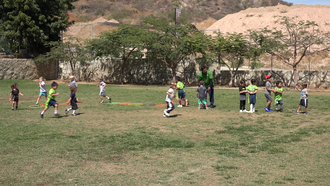 Children run on a grassy soccer field while a coach stands near training gear.