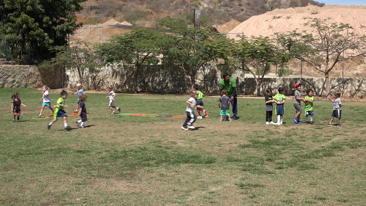 Several children run and walk across the San Jose del Cabo soccer field while the coach remains near the center group and training markers lie on the grass.