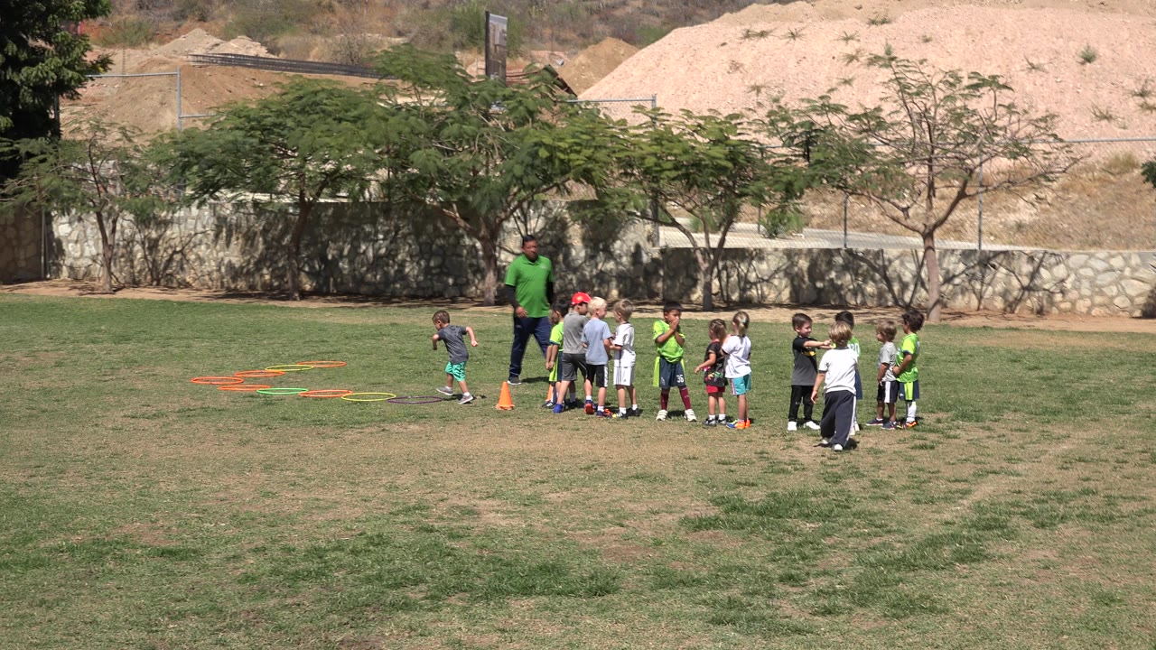 Children gather around a soccer coach beside cones and training rings on a grass field.