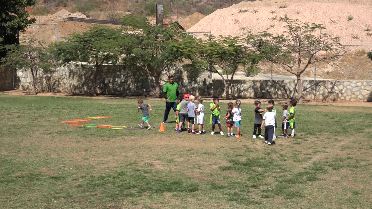 In San Jose del Cabo, one child moves left of the huddle near colored agility rings as the coach stands with the children on the grass.