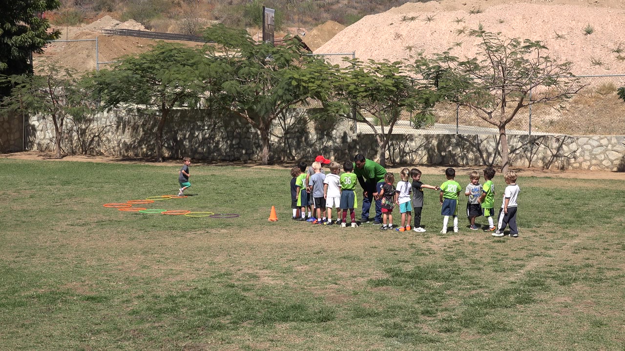 Children stand in a loose huddle with an adult coach on a grass field in San Jose Del Cabo, Mexico, with orange cones and agility ladders off to the left.