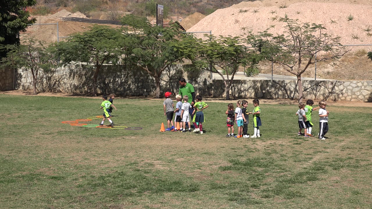 A young child runs through the drill area at left while the coach and other children wait together on the sunlit soccer field in San Jose del Cabo, Mexico.