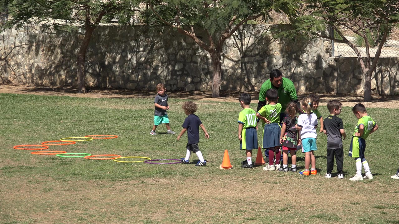 In San Jose Del Cabo, Mexico, young children gather around a coach on a grass field while colored hoops and an orange cone mark a training circuit at the left.