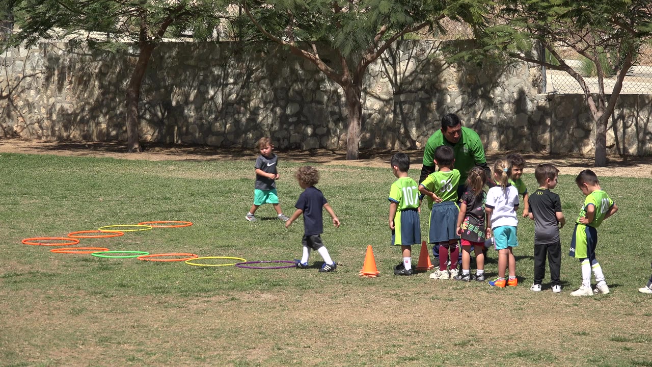 In San Jose Del Cabo, Mexico, one child moves near the training hoops while the coach bends toward a group of children beside orange cones on the grass.