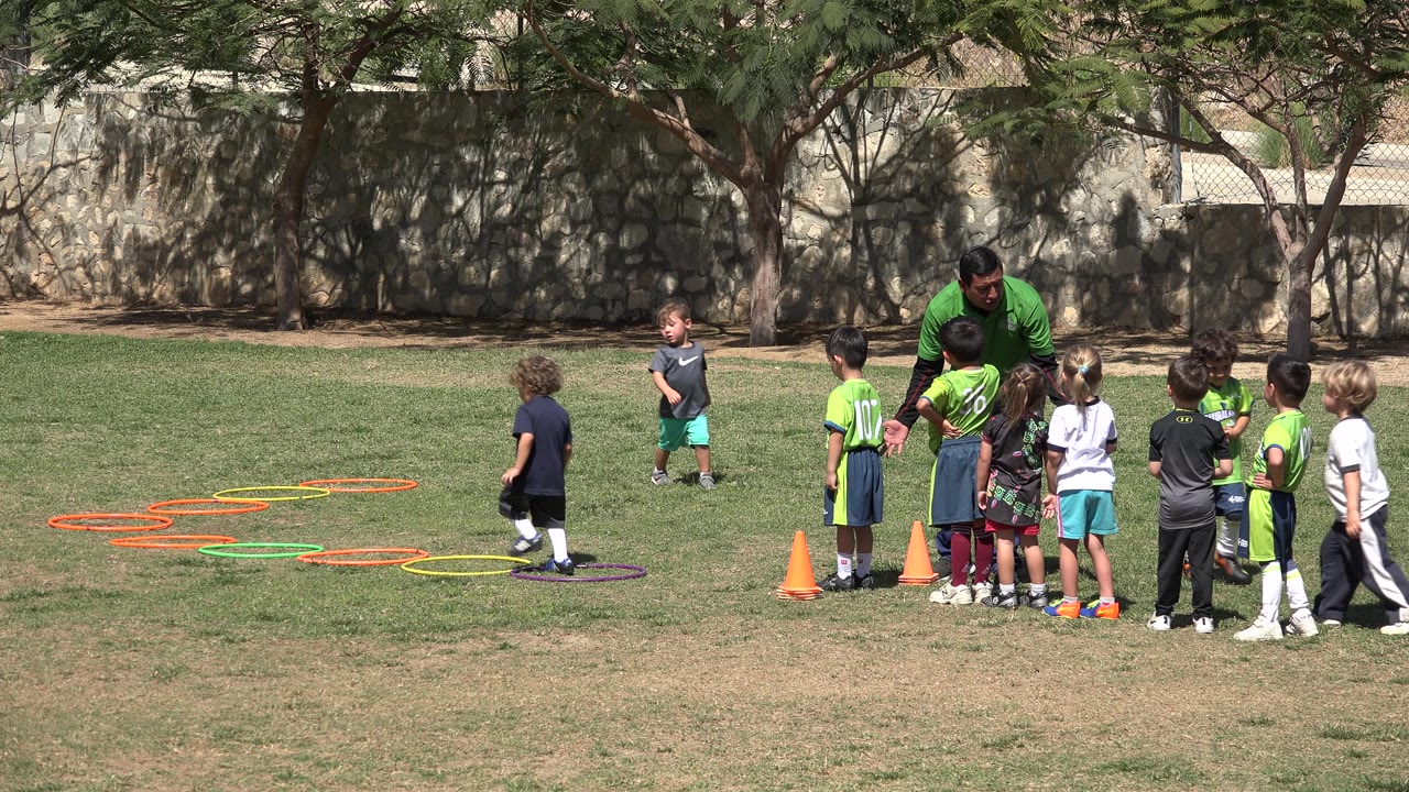 Children stand in a line near cones as a coach speaks to them.