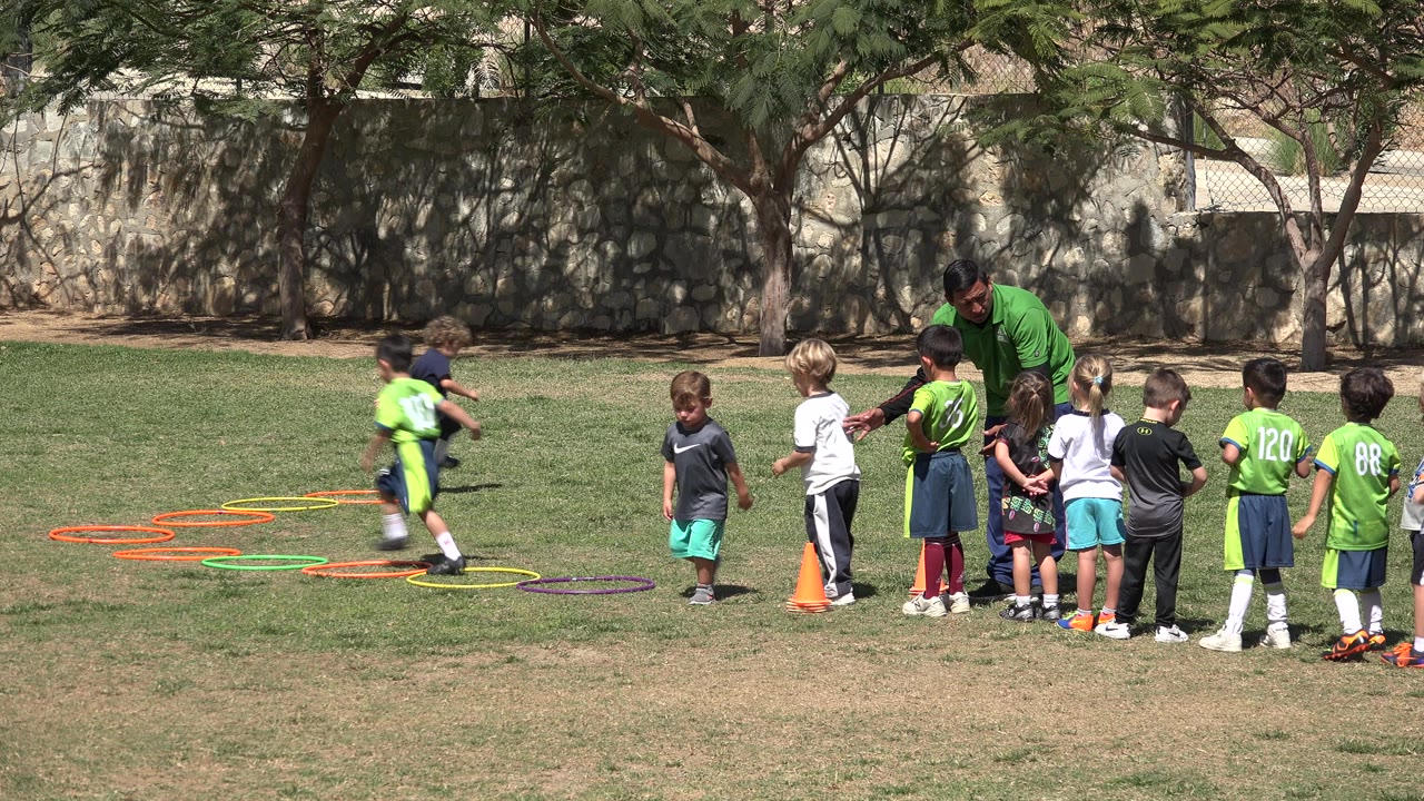 Children line up beside colored hoops and orange cones on a grass field.