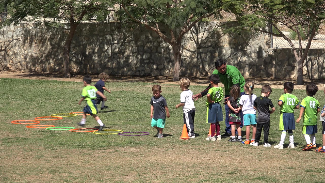 A child in a bright green shirt runs through colored hoops as classmates and a teacher wait nearby on a grass field in San Jose Del Cabo, Mexico.