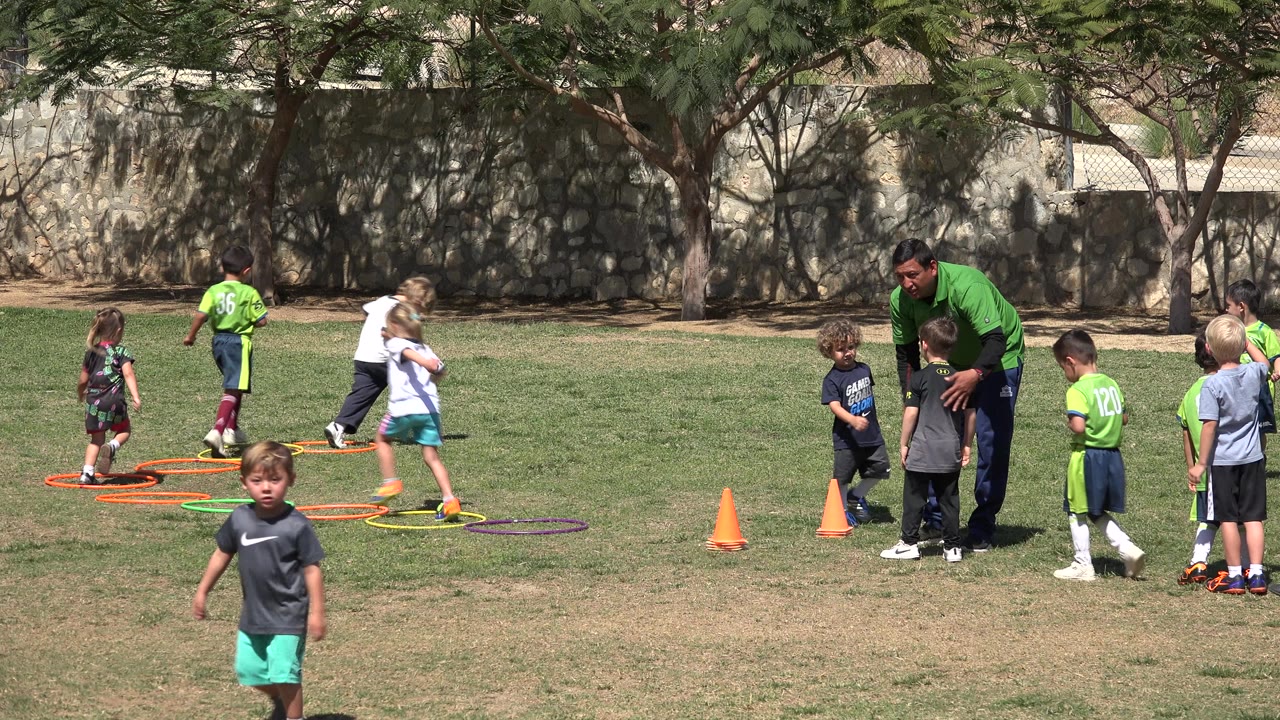 A child crosses the foreground while other children in green jerseys work near hoops and a coach watches on the field in San Jose Del Cabo, Mexico.