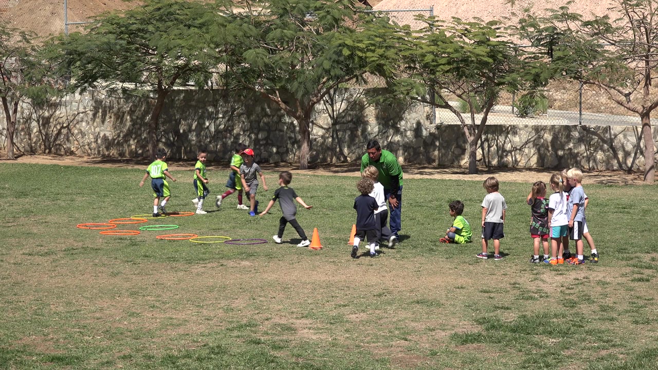 Children practice soccer drills on a grass field with a coach nearby.