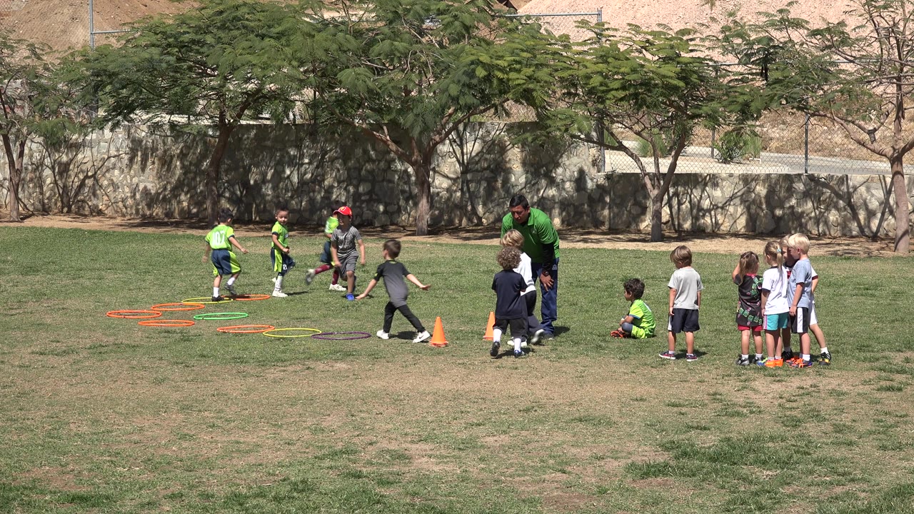 A child steps near the orange cones as other children wait in a small line and the coach watches from the right side of the field in San Jose del Cabo, Mexico.