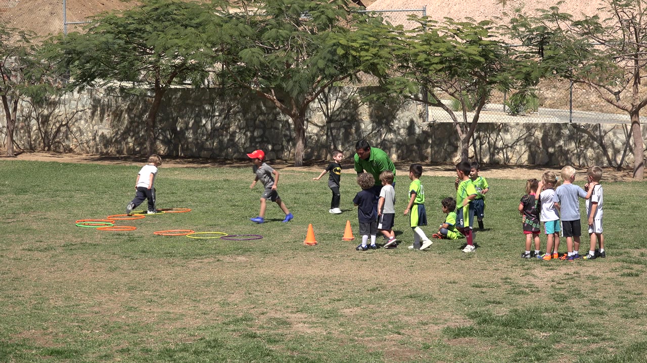 In San Jose del Cabo, Mexico, children cluster on a sunny grass field while an adult in green stands near colored hoops and orange cones at the start of the activity.