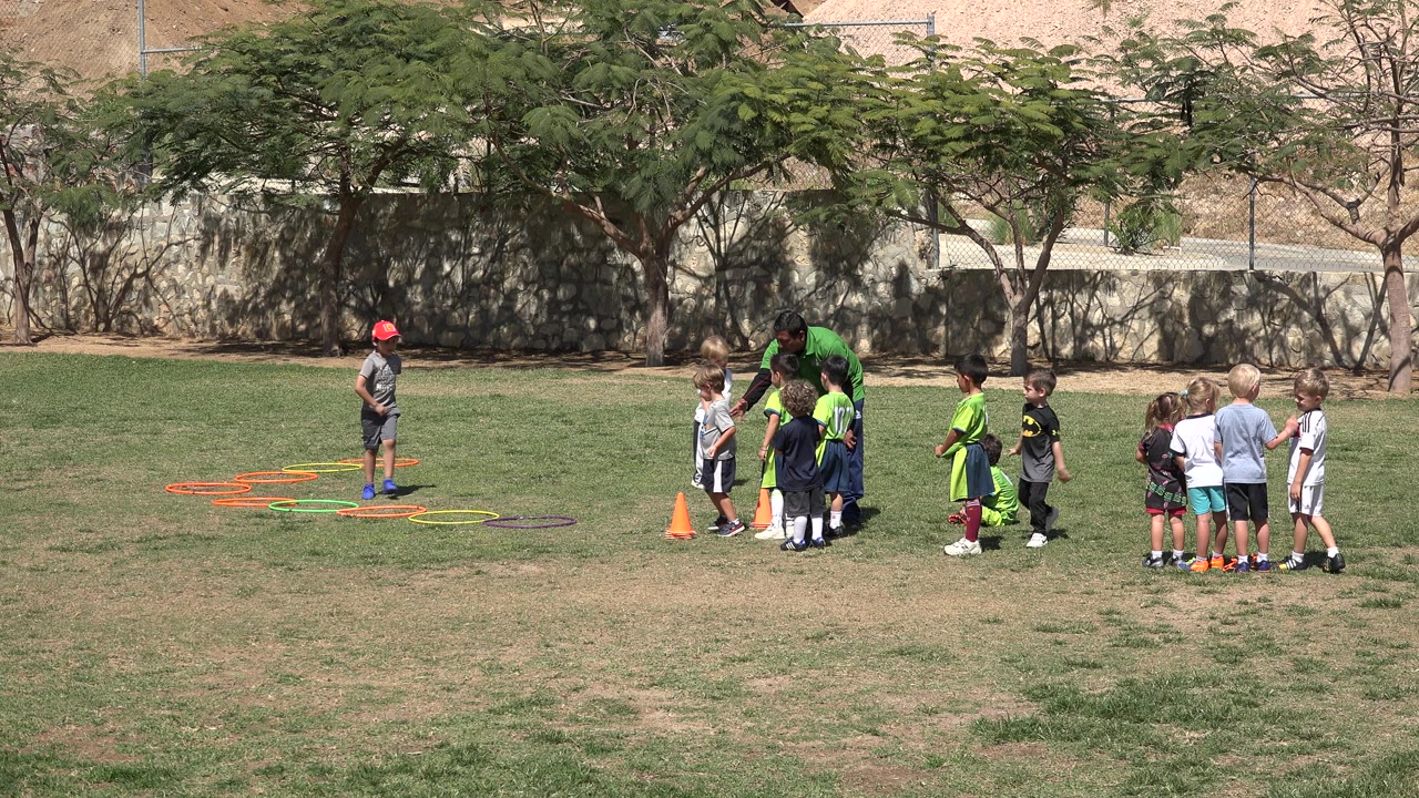 Children stand in small groups with a coach on a sunlit grass field in San Jose Del Cabo, Mexico, while orange cones and colored agility hoops sit nearby.