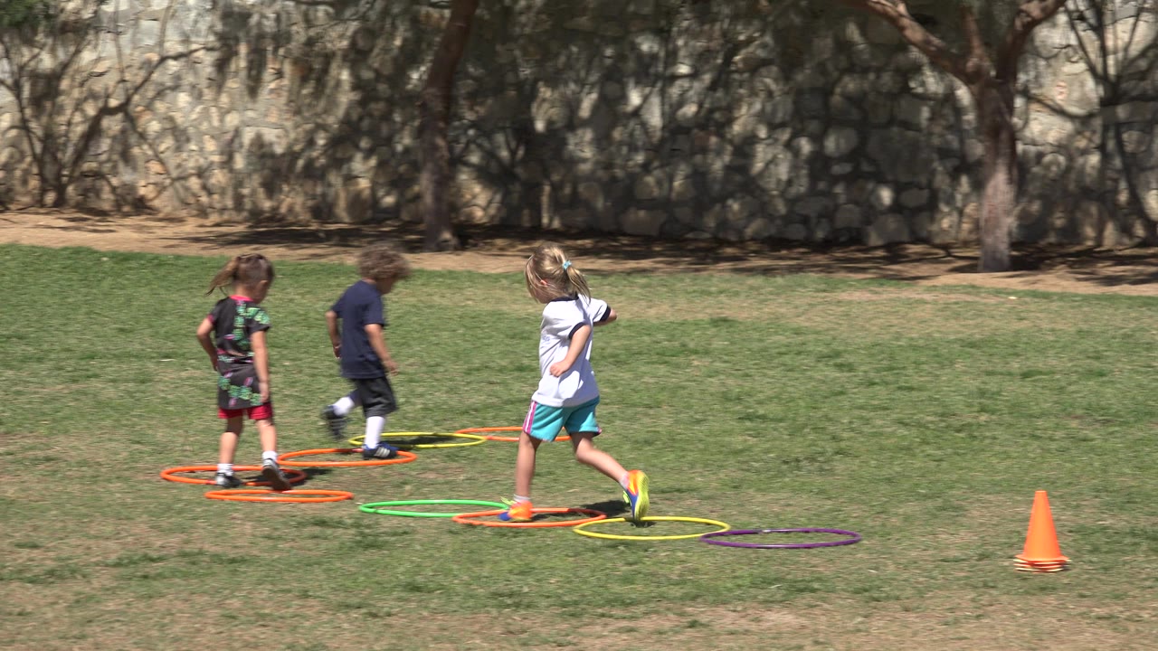 Three children run through orange, yellow, green, and purple hoops on a grass field in San Jose del Cabo, Mexico, with a stone wall and trees behind them.