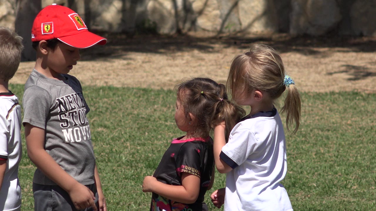 Three children meet on a sunny grass field in San Jose Del Cabo, Mexico, with a boy in a red cap facing two girls near the start of the clip.