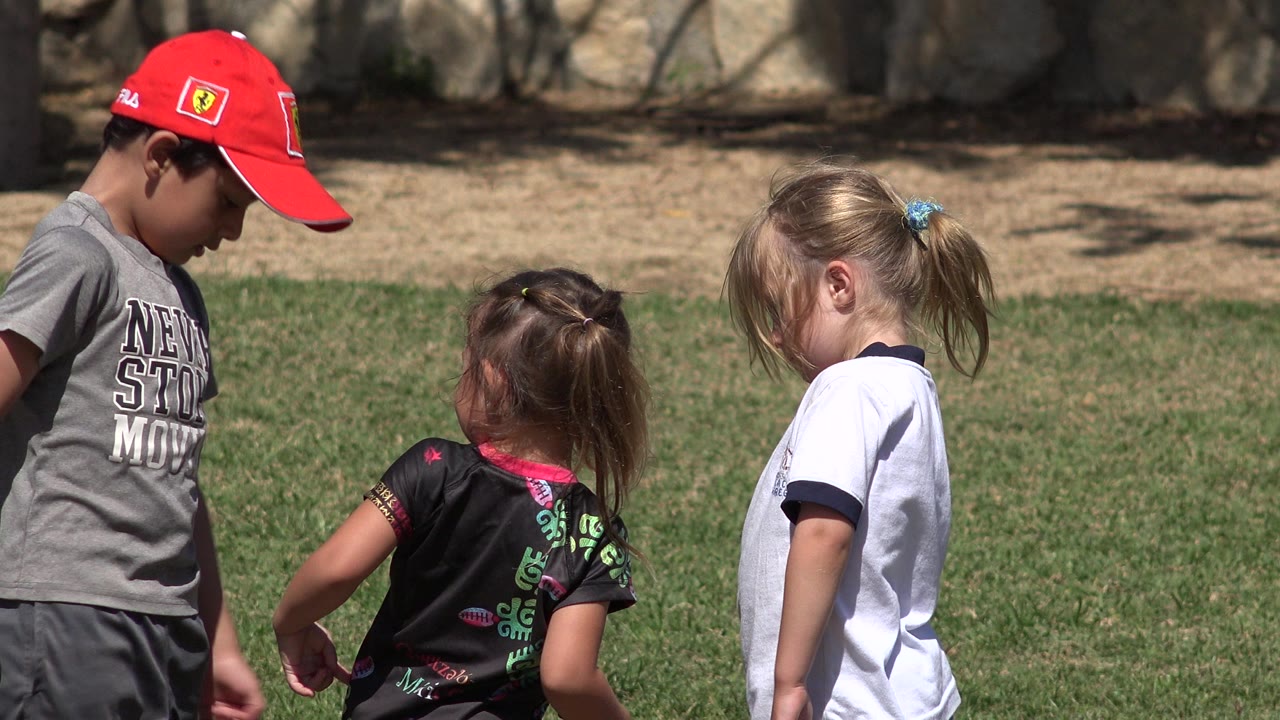 Three children stand close together on a grassy field in San Jose del Cabo, Mexico, with the boy in a red cap looking down near two girls.