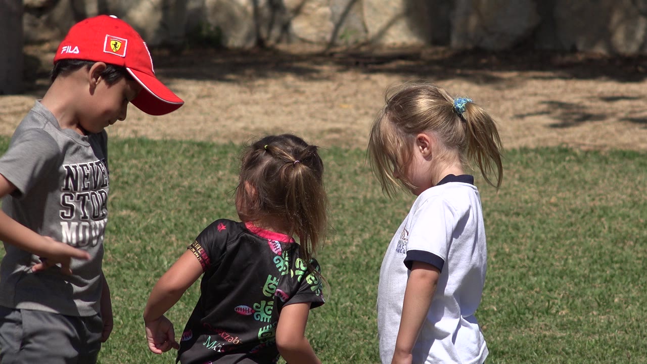 Boy in red cap looking down while two girls stand nearby on grass.