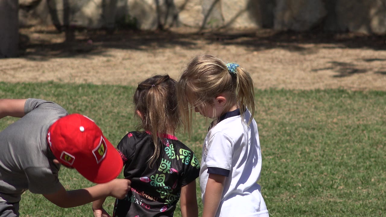 A boy in a red cap bends beside two girls on a grassy field in San Jose del Cabo, Mexico, with trees and a wall behind them.