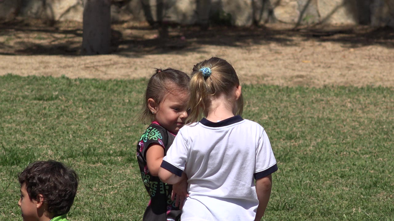 Two young girls stand close together on a grassy field in San Jose del Cabo, Mexico, while part of a boy is visible at the lower left edge.