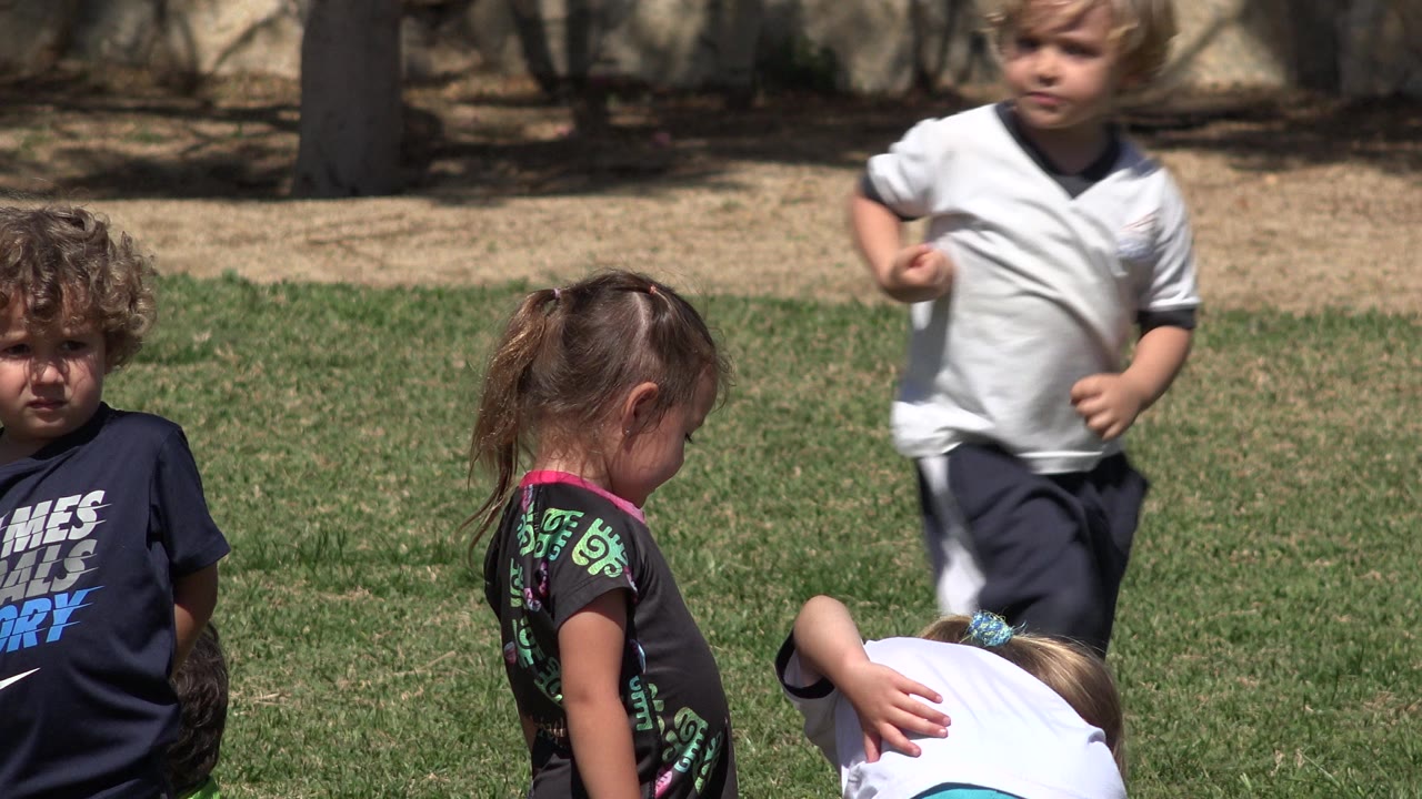 A curly-haired child in a navy shirt faces the camera while a girl stands turned away and a boy in a white shirt moves across the background on the San Jose del Cabo grass field.