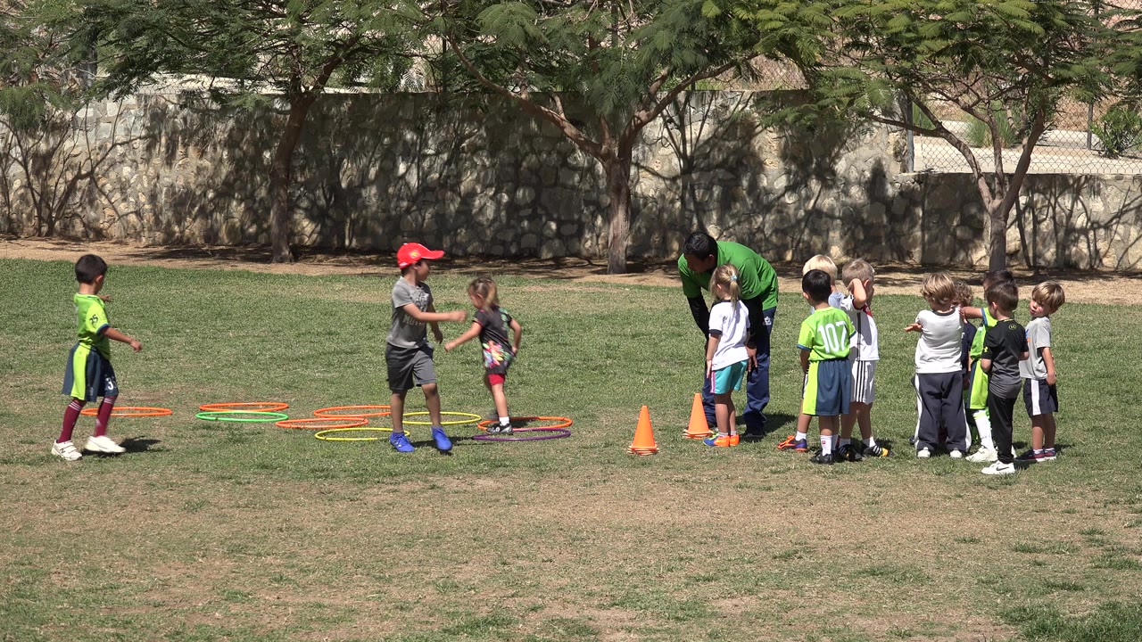 Children and a coach on a sunny grass field with cones and training hoops.