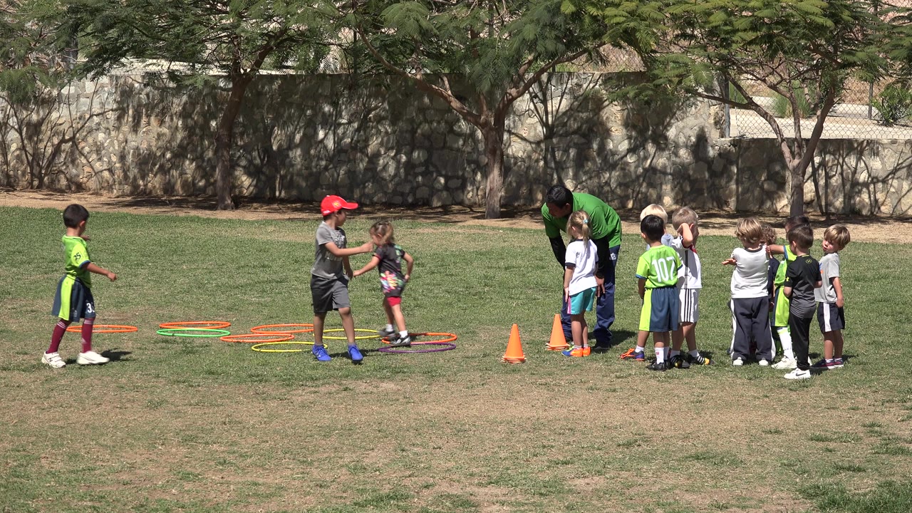 A child in a red cap stands near the colored hoops while other children cluster around the coach on the San Jose del Cabo park field.