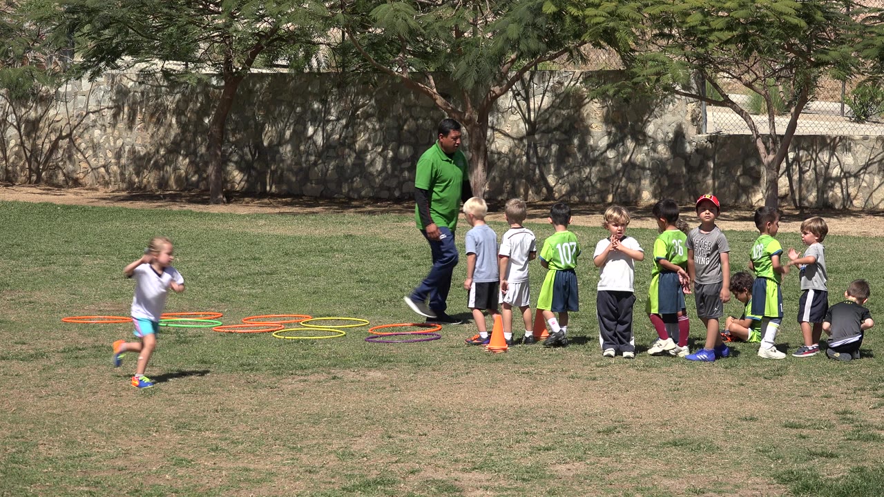 Children in athletic clothes gather beside orange cones and colorful hoops on a grass field in San Jose del Cabo, Mexico, while a coach stands near the line.