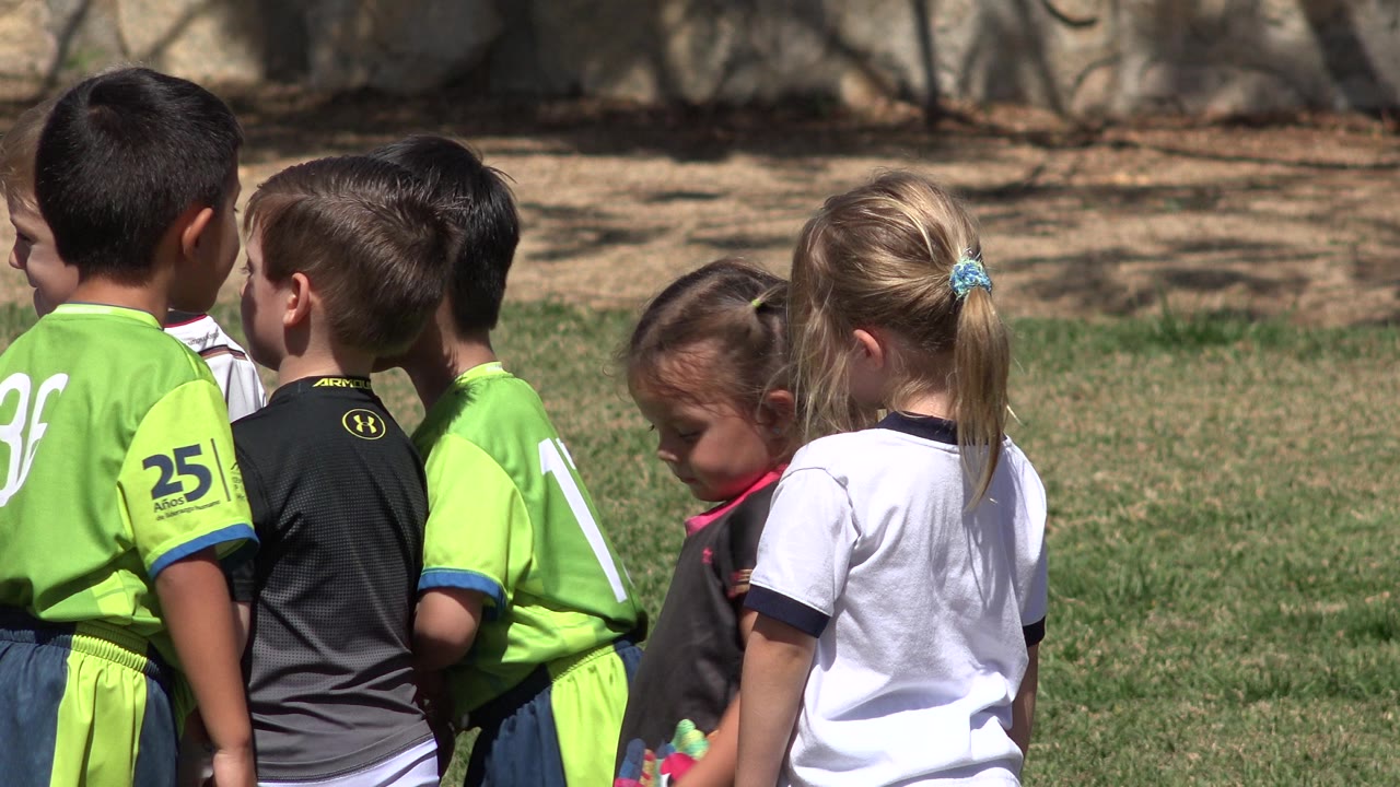 A close group of children in green, black, and white sports shirts stands on grass in San Jose del Cabo, Mexico, with one small child partly hidden near the center.