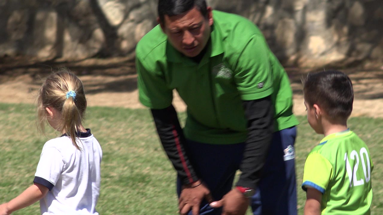 Adult man in a green shirt leans toward two children on a grass field in San Jose del Cabo, Mexico, with a stone wall and tree shade behind them.