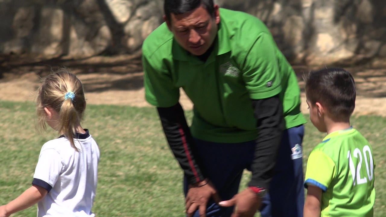 Adult trainer in green stands closer to the center of the frame while a girl in white and a boy in a green jersey listen on the grass field in San Jose del Cabo, Mexico.