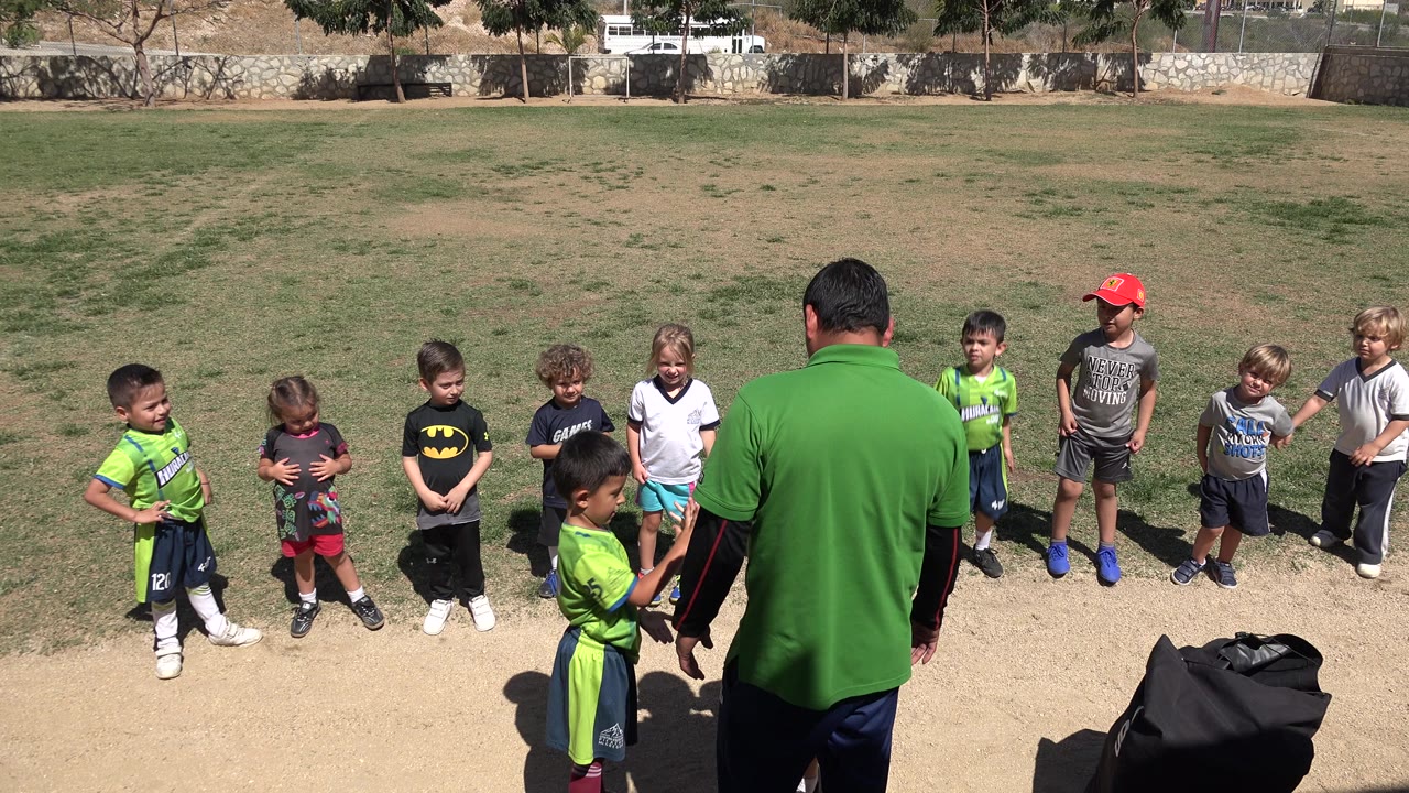 In San Jose del Cabo, Mexico, a soccer coach in a green shirt faces a small group of children standing on a sunlit grass and dirt field.