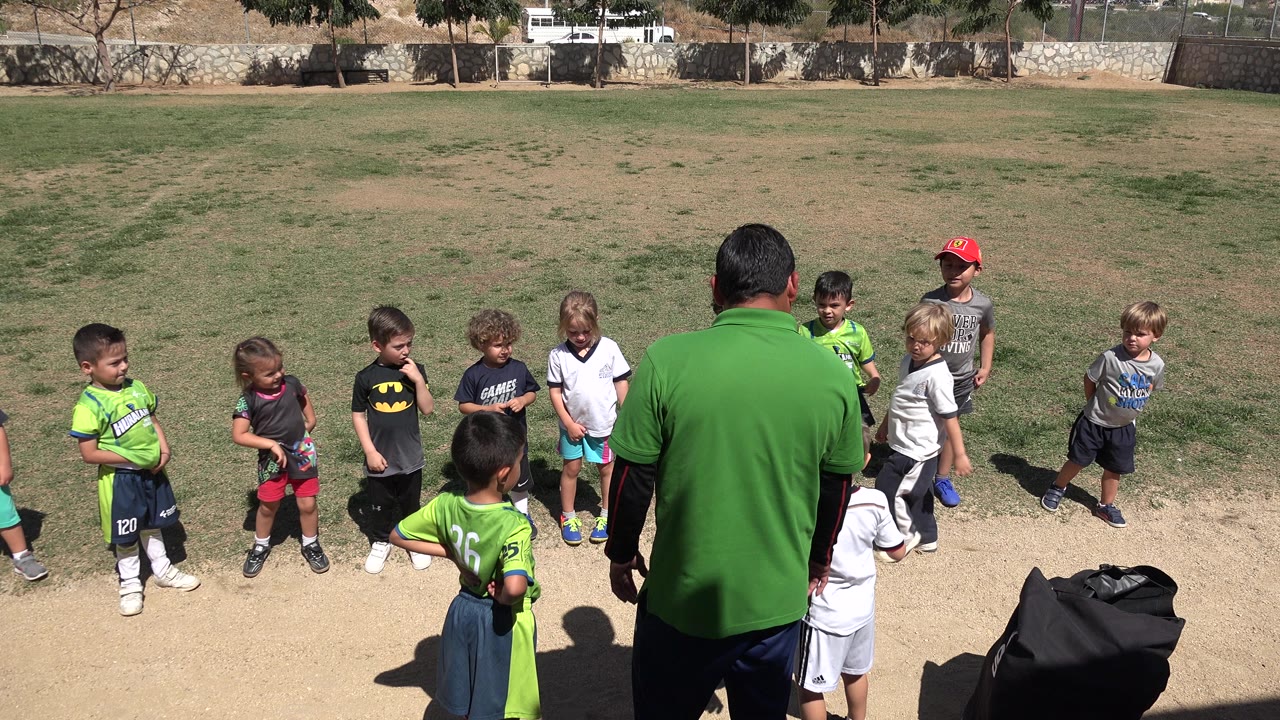 Children stand on a grass field around an adult supervisor in a green shirt.