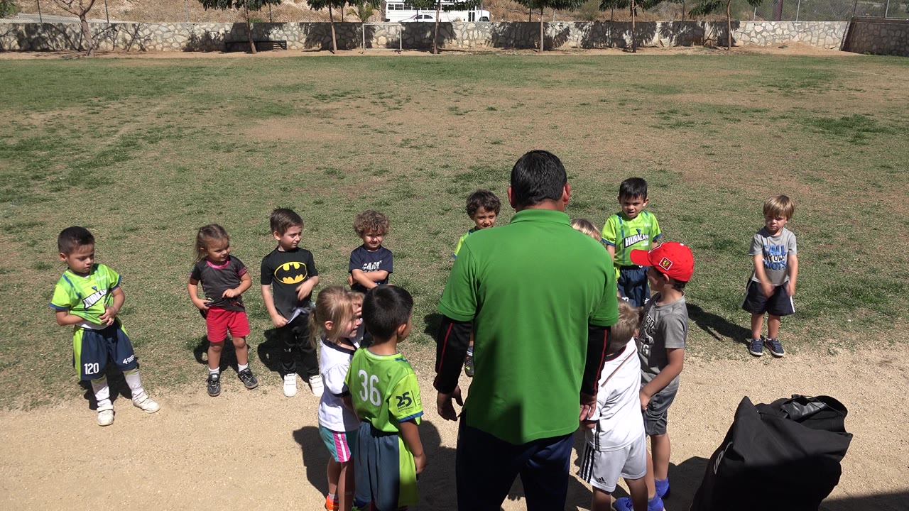At 0:00, young children in sports clothes gather around a man in a green shirt on an outdoor field in San Jose del Cabo, Mexico.