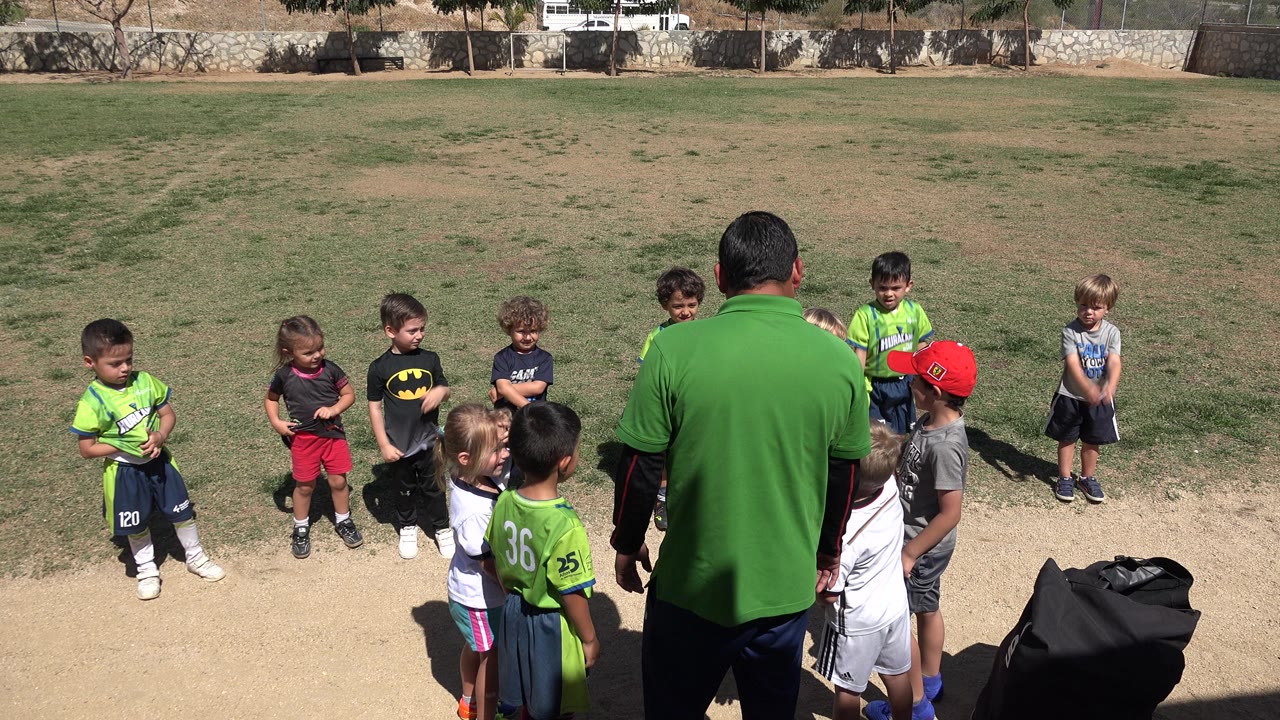 At 0:01, the man remains centered with his back to camera as children stand in a loose semicircle on the sunlit field in San Jose del Cabo, Mexico.