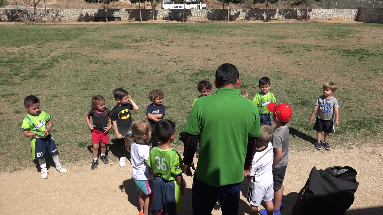 Children stand scattered near a man during outdoor soccer practice.