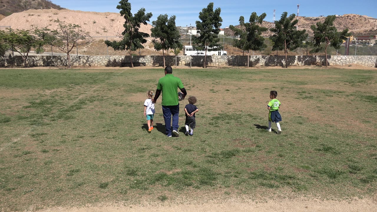An adult in a green shirt stands with three children on a patchy grass field in San Jose Del Cabo, Mexico, with trees, a wall, buildings, and tan hills behind them.