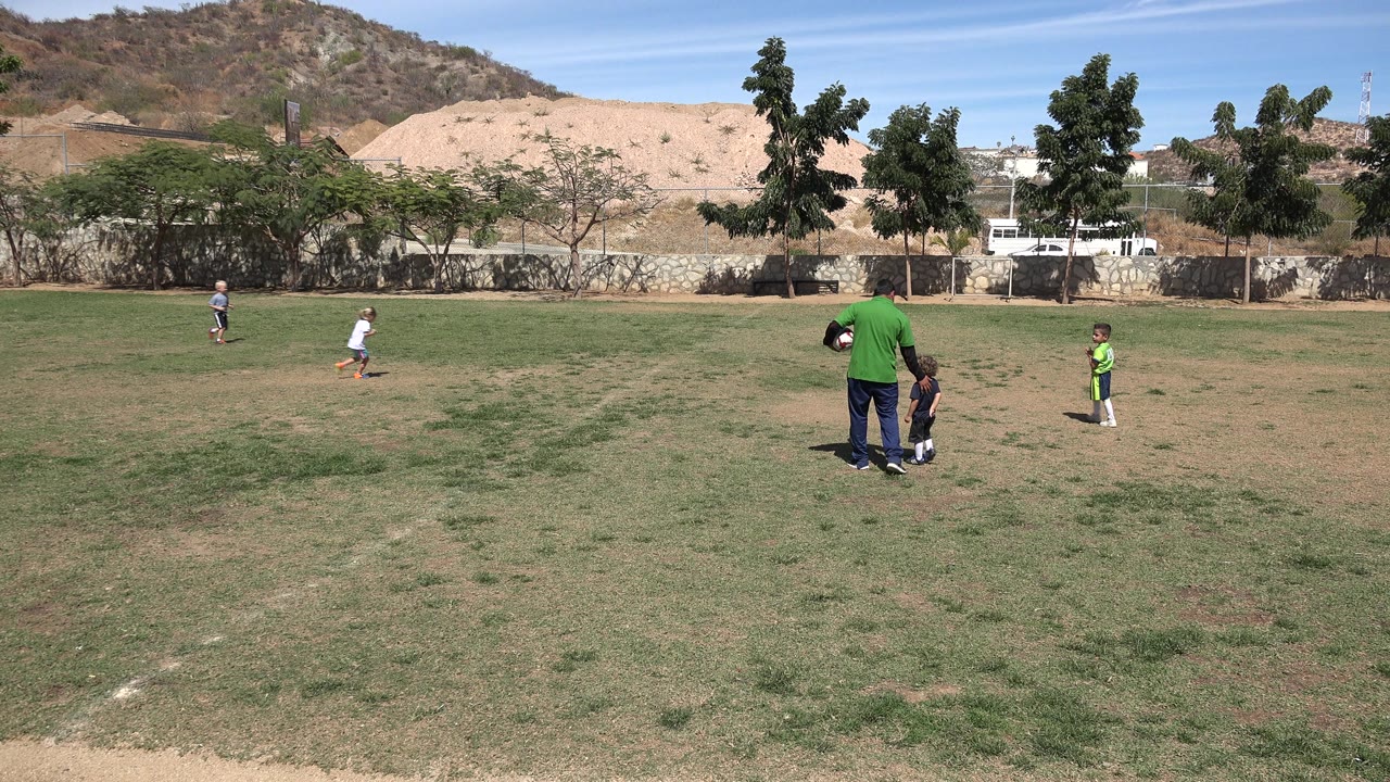 Wide view of a soccer field in San Jose Del Cabo, Mexico, with children spread across the grass and an adult standing near a child in the center.