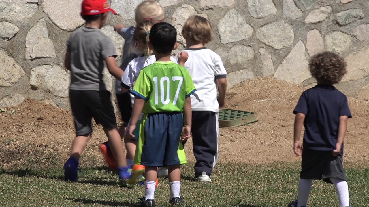 Children in soccer clothes stand with their backs to camera on a grass and dirt field in San Jose del Cabo, Mexico, with a stone wall behind them.