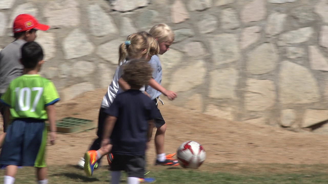 Children run after a soccer ball along a dirt edge.