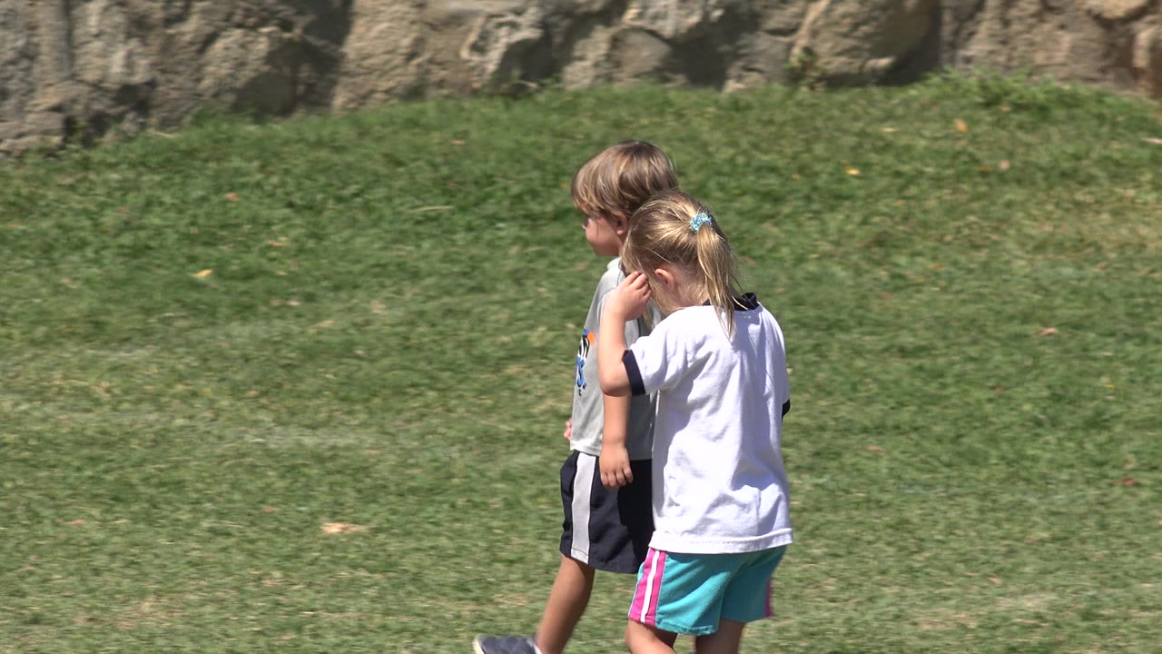 The girl in a white shirt walks close beside another child on the grass field in San Jose del Cabo, Mexico, with the stone wall filling the background.