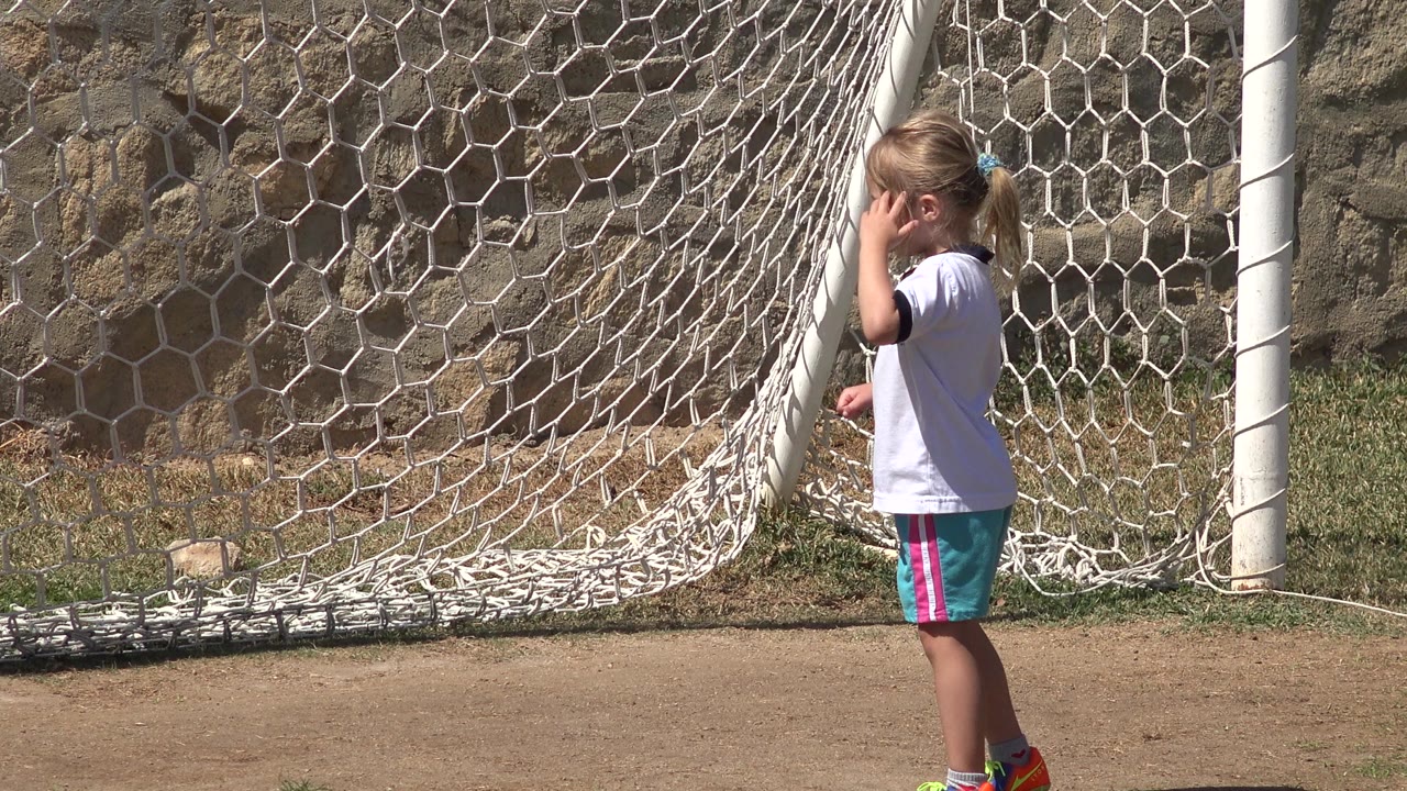 The girl remains beside the white goal net in San Jose Del Cabo, Mexico, with bright sun on the dirt field and stone wall.