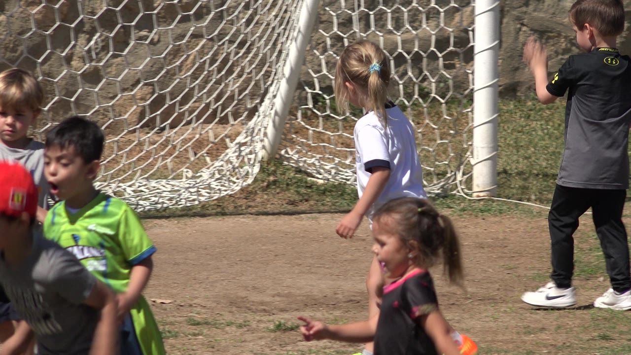 Children shift positions in front of the goal net in San Jose Del Cabo, Mexico, including a boy in a black shirt facing the net and younger kids crossing the foreground.