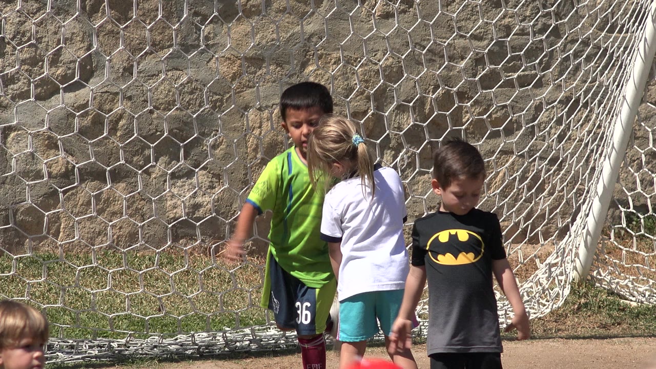 The children remain clustered in front of the soccer net in San Jose del Cabo, Mexico, with bright sunlight on their shirts and a stone wall filling the background.