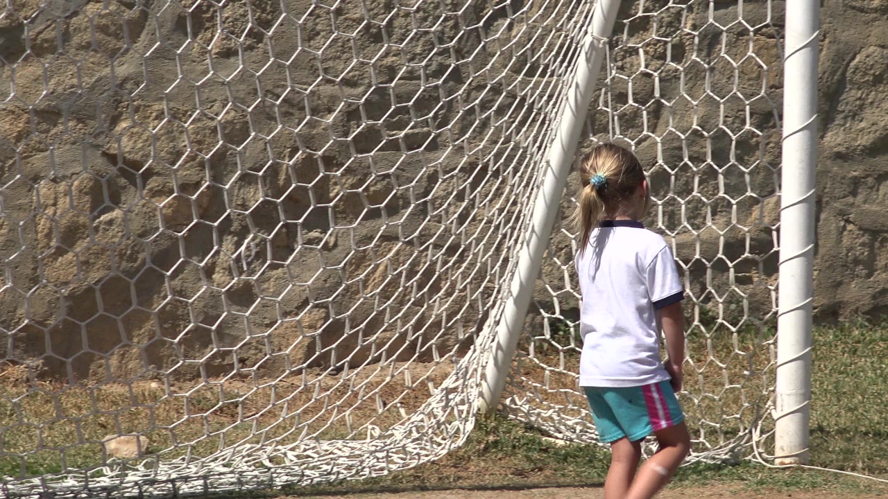 The girl is seen from behind walking beside the hanging soccer net, with grass underfoot and a stone wall in the background in San Jose del Cabo, Mexico.