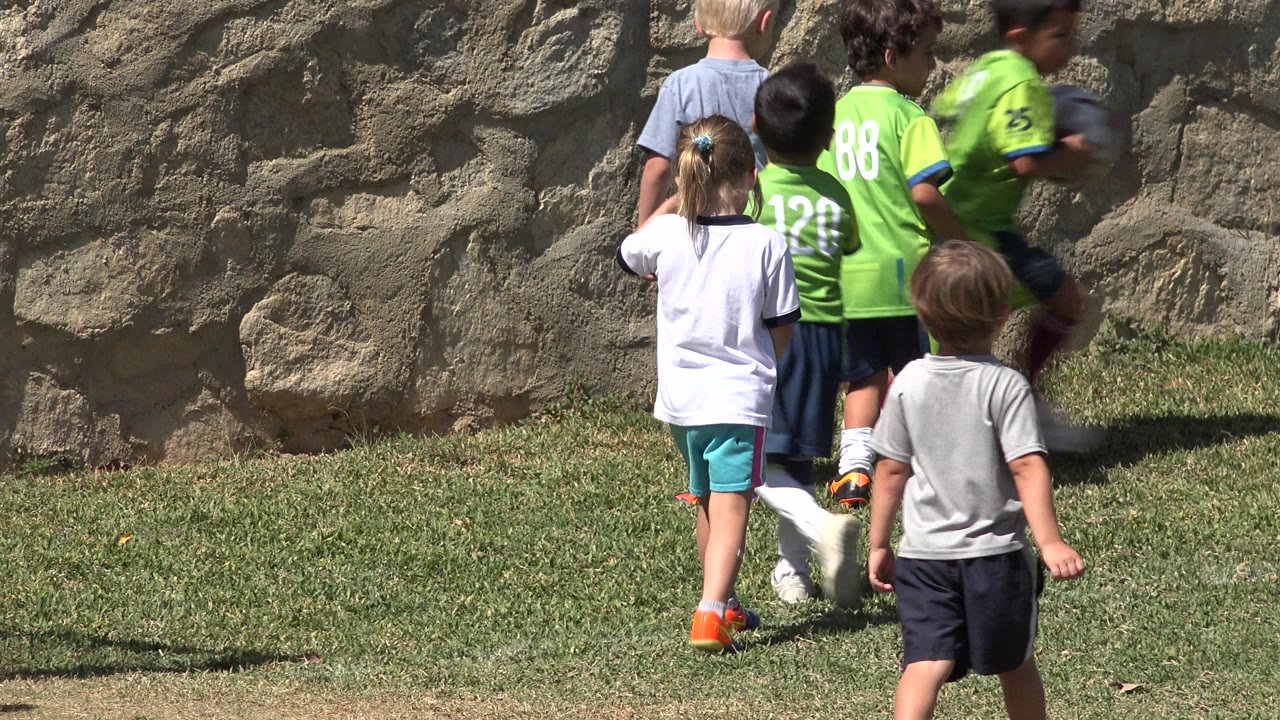 Several young children move across the grass in San Jose del Cabo, Mexico, with two lime green soccer jerseys and casual shirts visible against the stone wall.