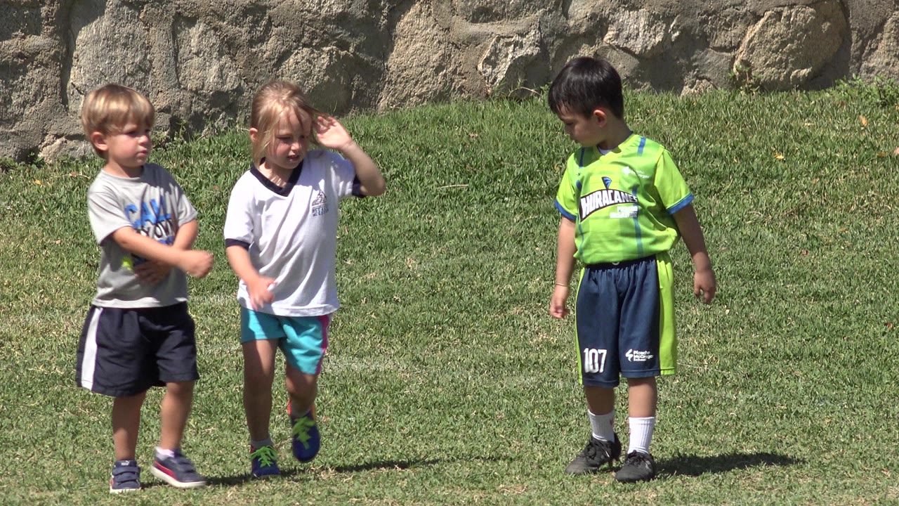 Three young children stand on a grassy park field in San Jose del Cabo, with one child in a bright green sports shirt and a rough stone wall behind them.