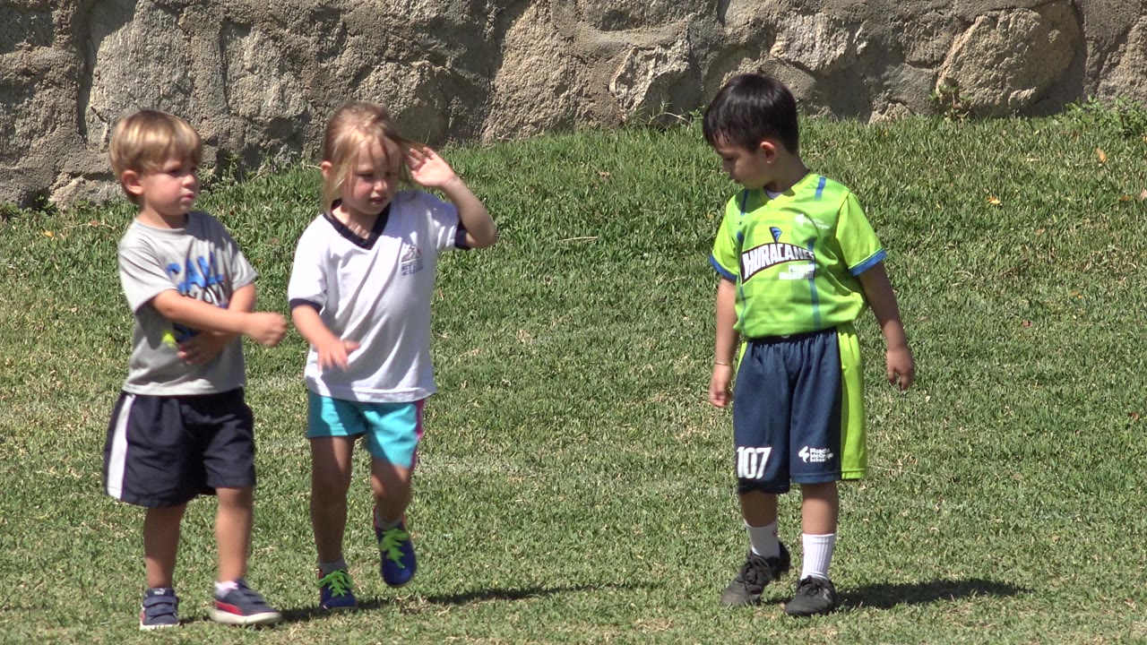 Three children remain in a loose line on the grass in San Jose del Cabo, with the girl in a white shirt lifting her hand near her face and the boy in green looking down.