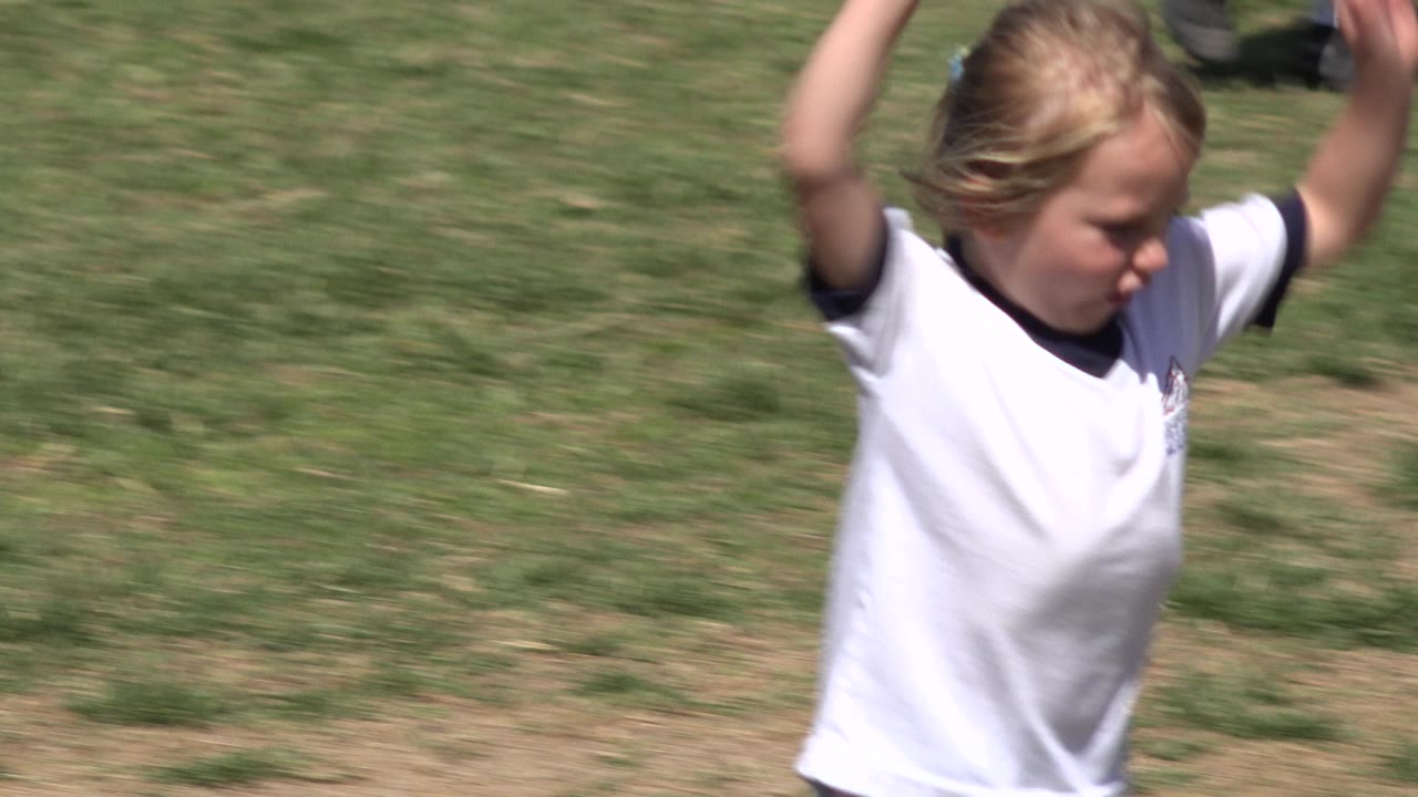 The child continues running with both arms raised, framed tightly against patchy green and brown grass in San Jose del Cabo, Mexico.