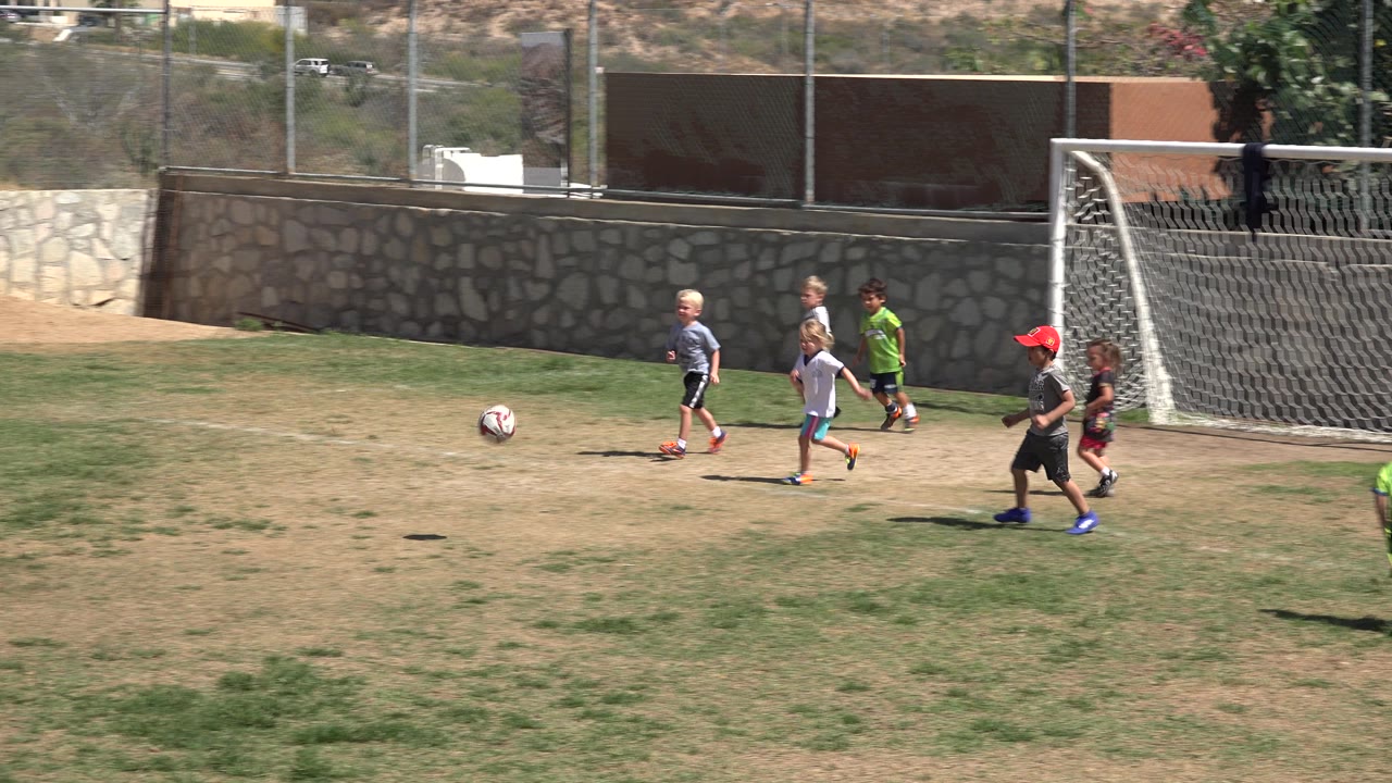 A soccer ball rolls across the field while several children run near the goal in San Jose del Cabo, Mexico, with the wall and fence still visible in the background.