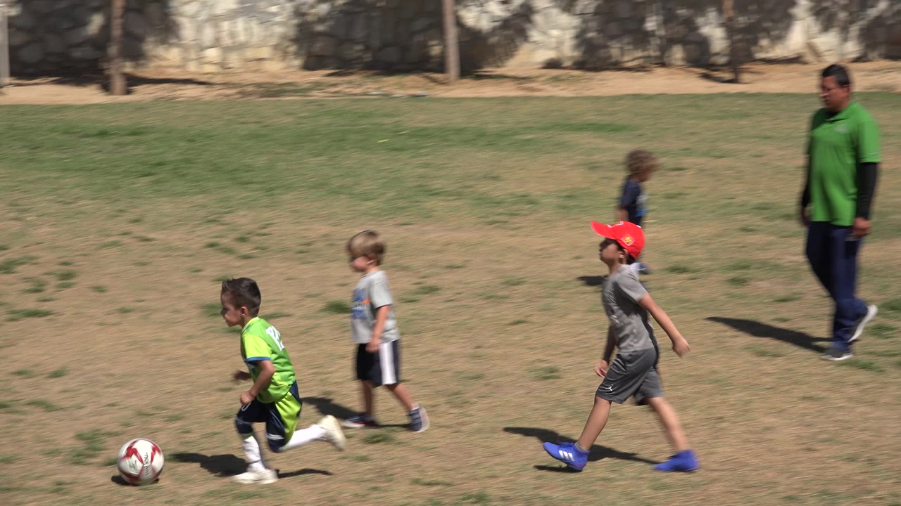 At 0:00 in San Jose Del Cabo, Mexico, young children run across patchy grass while a child in a bright green jersey moves near a red and white soccer ball.
