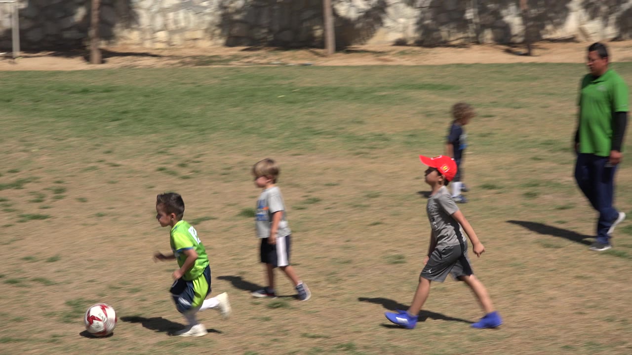 At 0:01 in San Jose Del Cabo, Mexico, the soccer ball sits ahead of a child in a green jersey while other children in gray shirts and a red cap move across the sunlit field.